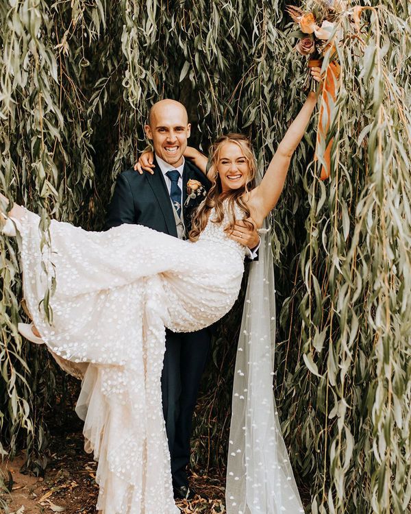 Groom in a navy suit lifts the bride up in front of willow tree