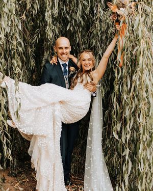 Groom in a navy suit lifts the bride up in front of willow tree