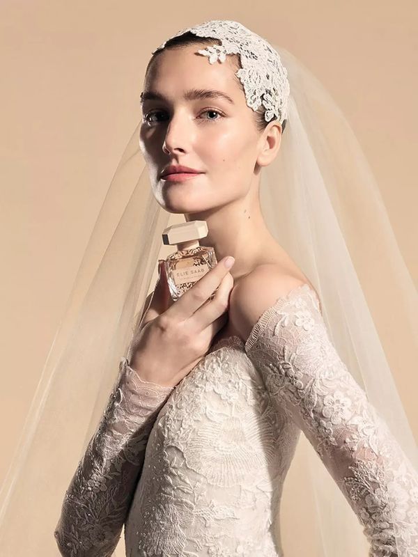 Bride wearing a lace wedding dress and veil, holding a bottle of Elie Saab Le Parfum Bridal Eau de Parfum Bridal Fragrance against a soft beige background - Wedding Perfume