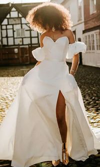 Bride wearing a modern off-the-shoulder wedding dress with puff sleeves and a thigh-high slit, standing on a cobbled street in natural sunlight - Wedding Dresses & Bridalwear