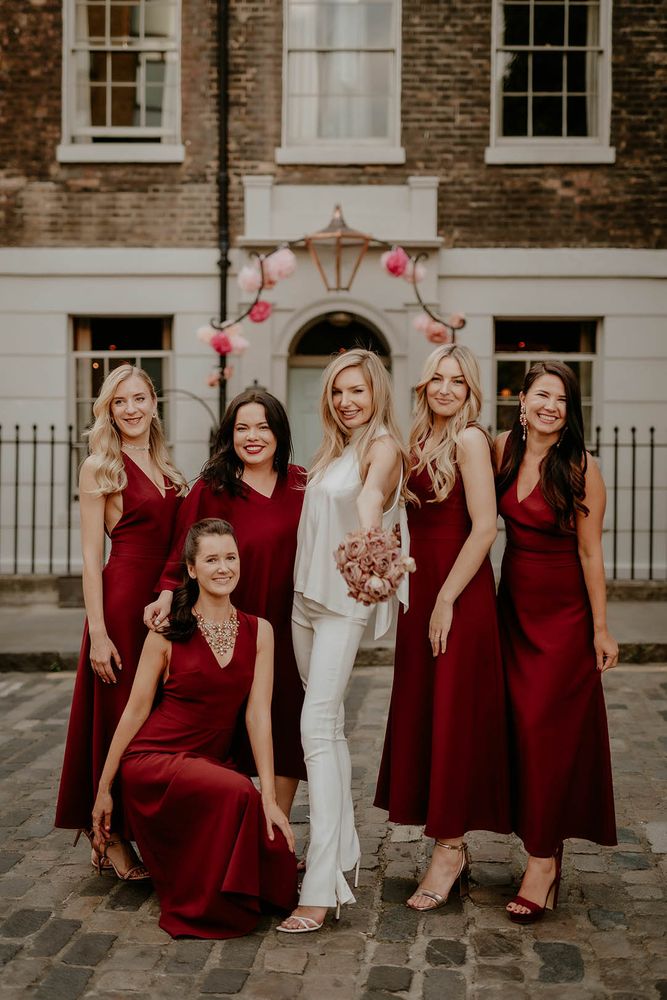 Bride wears high neck silk top complete with tapered and fitted white trousers beside bridesmaids in dark red bridesmaid dresses 