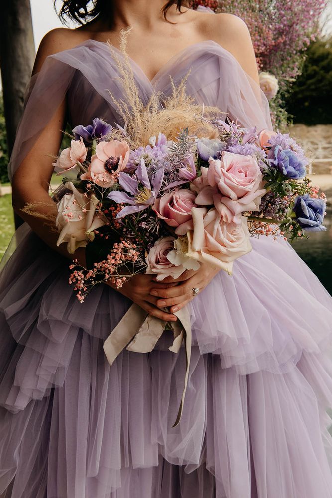 Bride wears off the shoulder purple tulle wedding dress complete with sweetheart neckline whilst carrying floral bouquet 