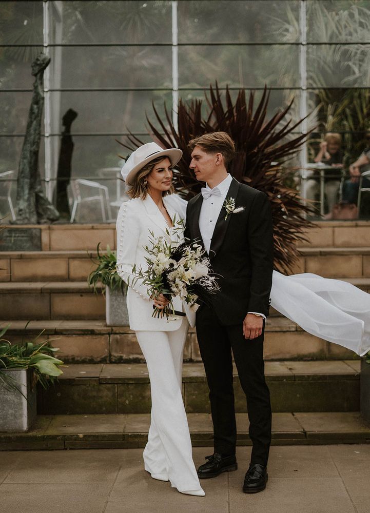 Bride wears Nadine Merabi bridal suit with pearl embellishment and white bridal hat complete with white floral bouquet 