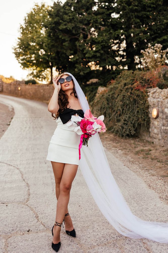 Bride wears short wedding dress with statement black bow to front and colourful pink floral bouquet