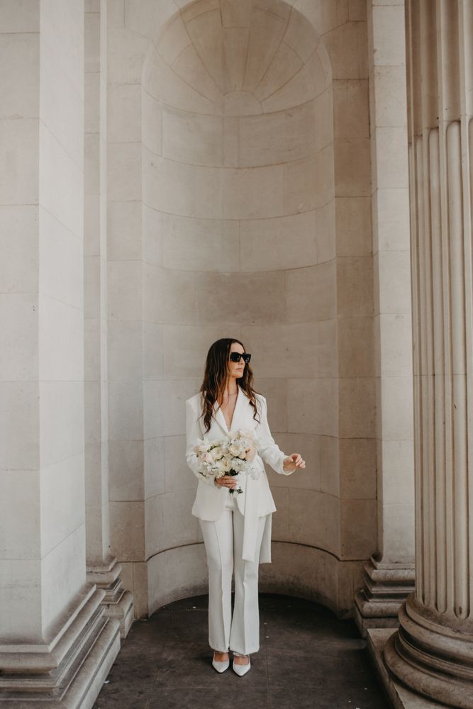 Bride wears fitted bridal suit with tie to waist and tapered trousers with flared bottoms 