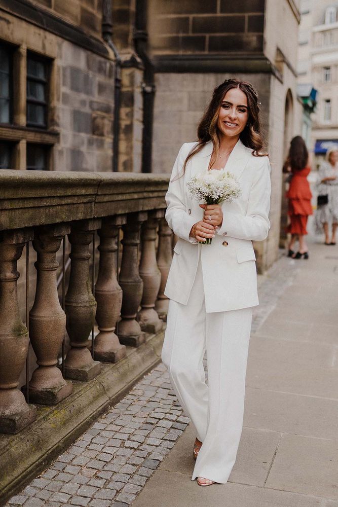 Bride wears white suit jacket complete with wide leg trousers and white rose bouquet
