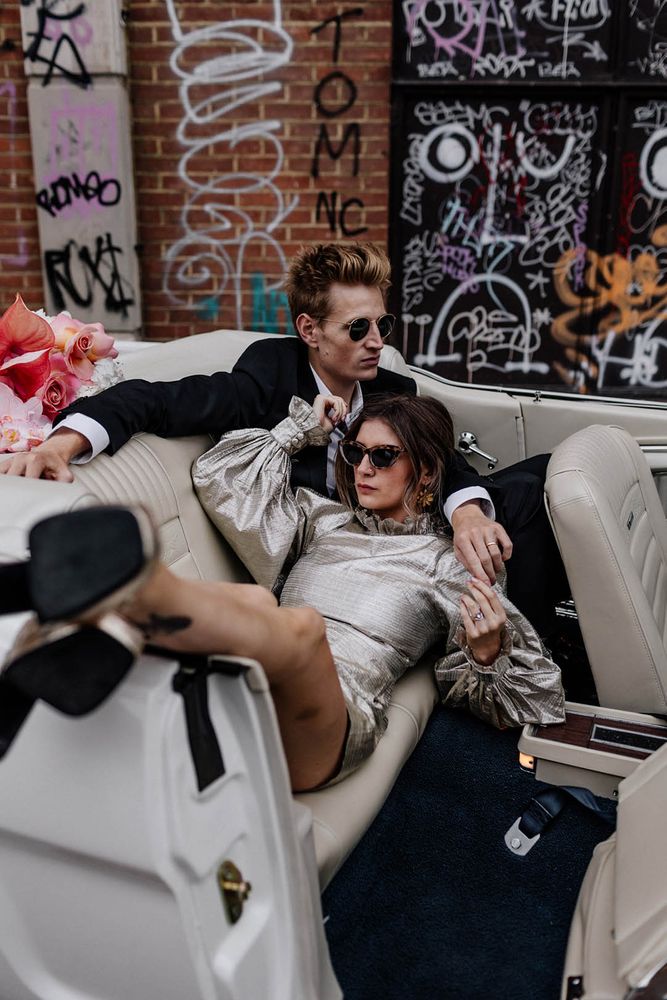 Bride wears sunglasses and metallic short wedding dress complete with puffed long sleeves and sits in car with her husband 