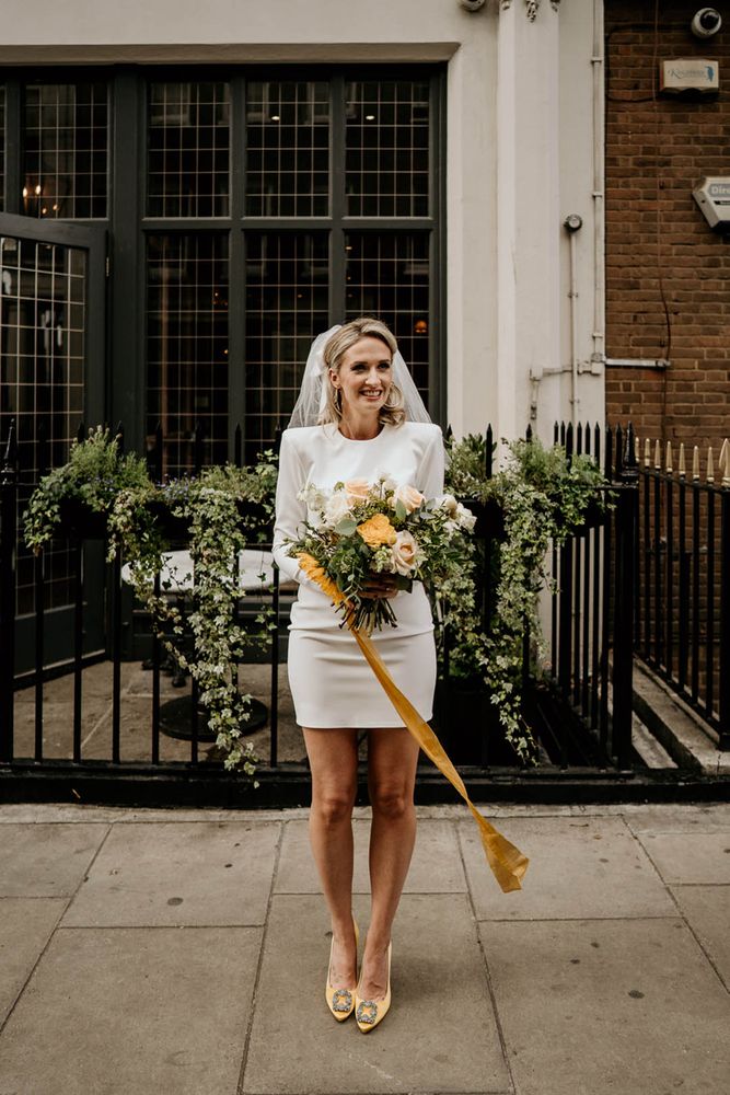 Bride in a short wedding dress with long sleeves and shoulder pads and yellow Manolo Blahnik pumps holding a sunflower bouquet 