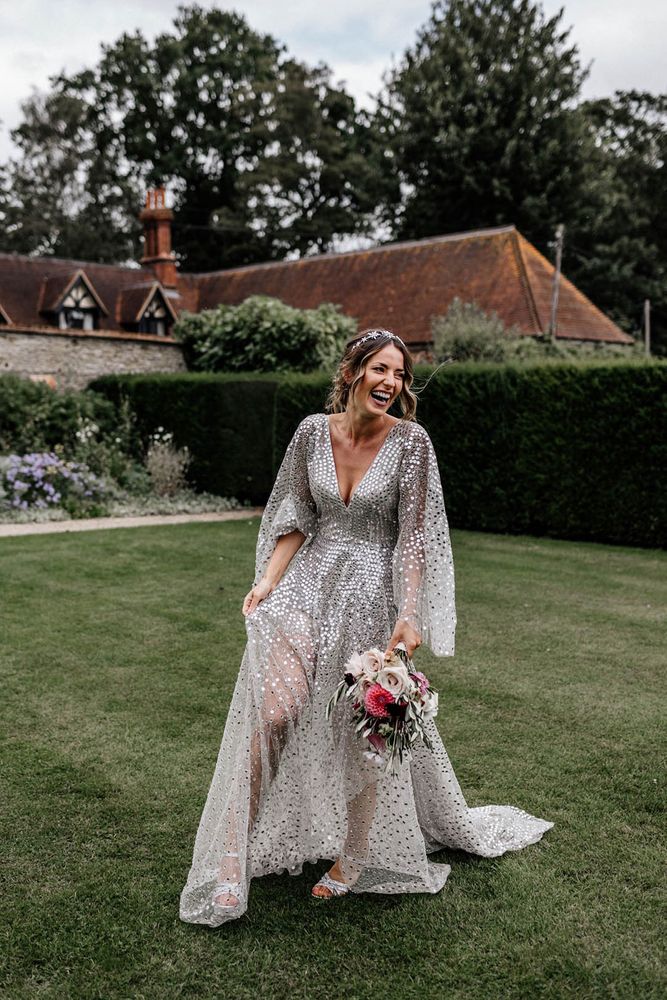 Bride wears silver sequin wedding dress with long sheer full sleeves and bridal crown whilst holding colourful floral bouquet 