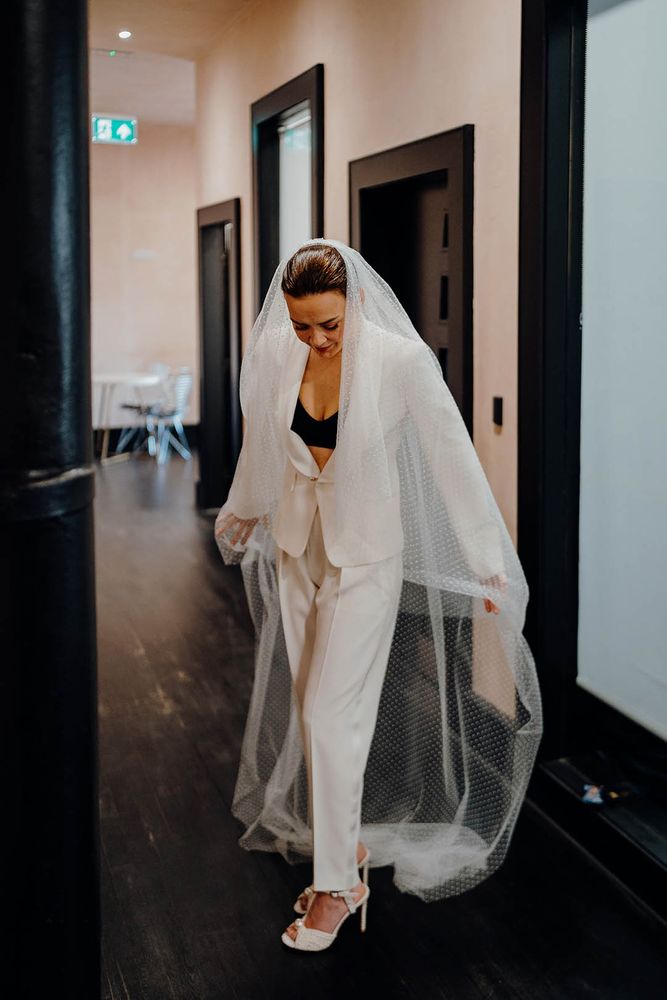 Bride wears white bridal suit with black bandeau top beneath and floor length veil 