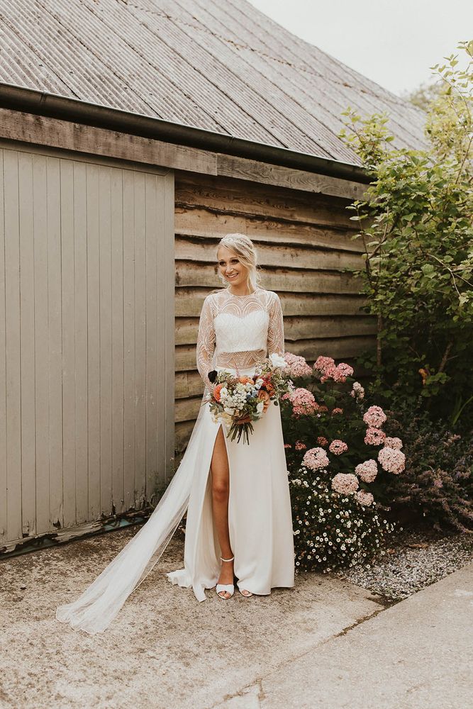 Bride in Catherine Deane wedding dress separates with lace top and side slit skirt holding an orange dahlia wedding bouquet 